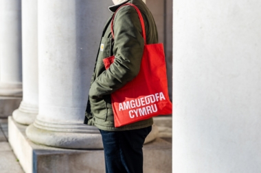 A red Amgueddfa Cymru / Museum Wales tote bag worn over the shoulder of a male model.